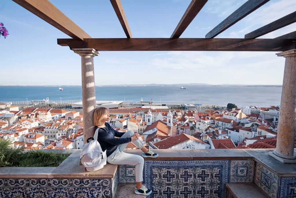Miradouro em Alfama com mulher sentada em mureta decorada com azulejos azuis e vista dos telhados vermelhos e do rio Tejo, típico passeio para adolescentes em Lisboa que gostam de miradouros e boas fotos | 10 melhores passeios para adolescentes em Lisboa #3