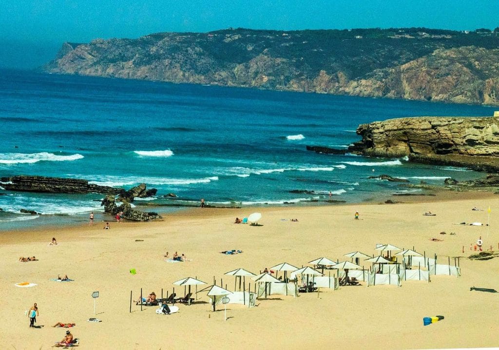 Praia de Carcavelos com areia clara, mar azul, ondas suaves e algumas pessoas tomando sol e caminhando perto da água, local muito procurado por adolescentes em Lisboa para surf e esportes ao ar livre | 10 melhores passeios para adolescentes em Lisboa #6